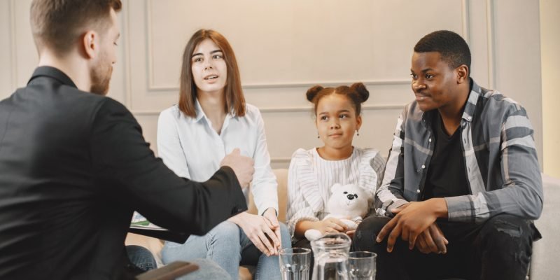 Family counseling session at home with therapist. Pshycologist showing pictures of emotions to a girL.African American father and European mother.